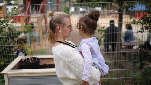 Eine jüngere Frau mit Brille hält ein Kleinkind im Arm und spricht mit ihm. Im Hintergrund steht ein Hochbeet.