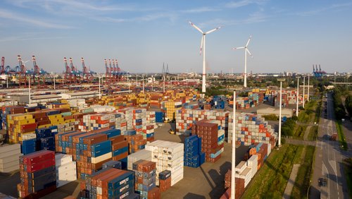 Viele, gestapelte Container im Hafen von Hamburg. Rechts im Bild Windräder und eine Straße.