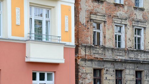 Eine frisch renovierte Altbaufassade steht in leuchtenden Farben neben einer verfallenen unrenovierten, die noch sichtbare Kugellöcher aus dem zweiten Weltkrieg trägt.