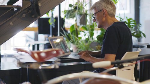Nachhaltiges Hot Desking - zeigt die Hände eines reifen Mannes, der an einem Laptop auf einem Schreibtisch voller Zimmerpflanzen mit einem Retro-Fahrrad im Vordergrund arbeitet.