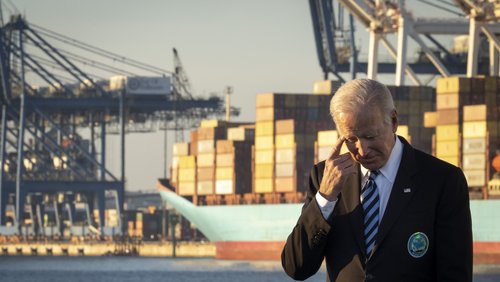US-Präsident Joe Biden steht vor dem Panorama eines Hafenterminals am Hafenbecken. Er blickt nachdenklich nach unten und greift an seine Schläfe..