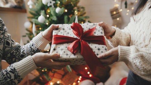 Couple exchanging christmas gift with red bow on background of christmas tree with lights. Stylish couple hands holding present with red ribbon close up in festive decorated room. Happy Holidays Couple exchanging christmas gift with red bow on background of christmas tree with lights. Stylish couple hands holding present with red ribbon close up in festive decorated room. Happy Holidays