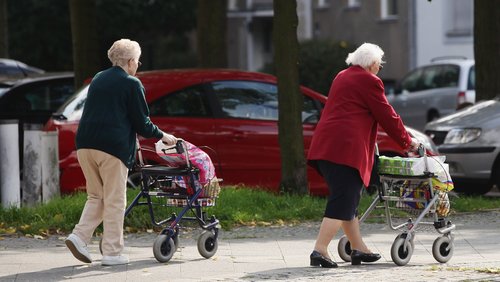 Zwei ältere Damen gehen mit Rollatoren eine Straße entlang.
