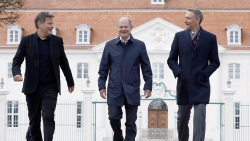 Robert Habeck, Olaf Scholz und Christian Lindner gehen vor Schloss Meseberg auf die Kamera zu; sie unterhalten sich und lachen.