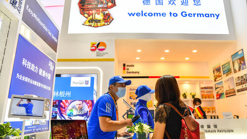 Ein Mann berät eine Frau an einem Messestand in China. Über dem Stand steht auf einem Schild "Welcome to Germany". Es sind deutsche und chinesische Flaggen zu sehen. Auf verschiedenen Werbeflächen werben Unternehmen und Regionen in chinesischen und lateinischen Schriftzeichen.