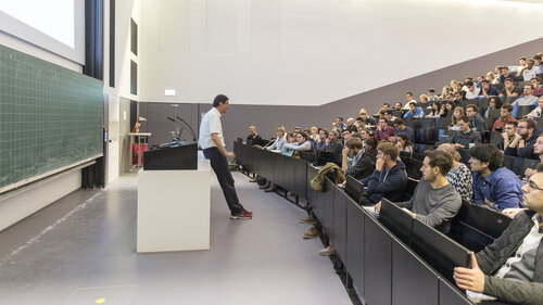 Ein Hörsaal. Ein Professor lehnt an seinem Pult und spricht zu einem vollbesetzten Hörsaal.