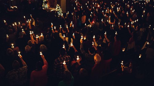 In einer dunklen, weihnachtlich geschmückten Kirche stehen viele Menschen in den Bankreihen. Sie halten Kerzen in die Luft, die den Raum in ein schummriges Licht tauchen.