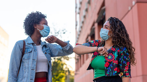 Young and friendly african girl and caucasian girl keeping social distance, greeting each other by bumping elbows instead of hugs, kisses or handshaking, wearing mask for prevent coronavirus infection