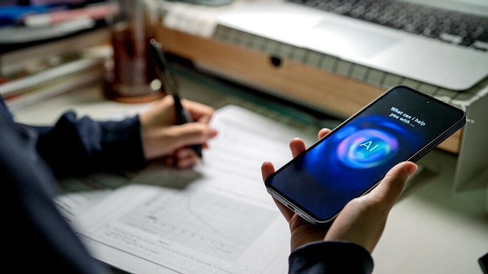 Ein Student kommuniziert über sein Smartphone mit einem KI-Chatbot, während er an einem Schreibtisch mit Laptop, Notizen und Schreibutensilien lernt. Ein Student kommuniziert über sein Smartphone mit einem KI-Chatbot, während er an einem Schreibtisch mit Laptop, Notizen und Schreibutensilien lernt.