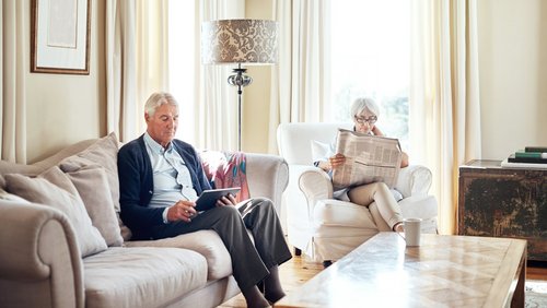 Aufnahme eines älteren Mannes, der ein digitales Tablet benutzt, während seine Frau zu Hause eine Zeitung liest