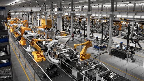 In einer hell erleuchteten, klinisch aussehenden Fabrikhalle fertigen Roboterarme einen langen Strom an Autokarosserien.