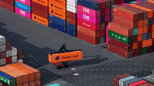 Ansicht von oben. Ein Fahrzeug bewegt einen Schiffscontainer durch ein Hafengelände.