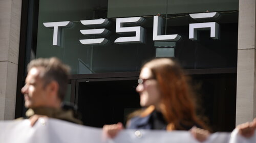 Im Hintergrund ist das Logo einer Tesla-Filiale zu sehen, davor befinden sich Demonstranten.