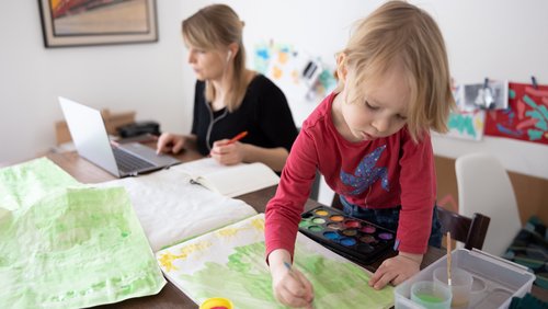 Ein Kind malt mit Wasserfarben, während seine Mutter am Tisch neben ihm vor einem Computer sitzt.