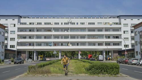Weitwinkel: Ein weißes langgezogenes Mehrfamilienhaus unter dem eine Straße verläuft. In der Mitte der Straße ein Grünstreifen, darauf ein Mann mit Hund.