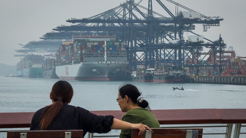 Zwei Chinesinnen sitzen auf einer Bank und blicken auf ein Hafenterminal. Dort sind große Kräne zu sehen, die mehrere Containerschiffe beladen.