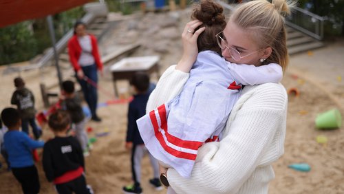 Eine Erzieherin hält ein kleines Kind auf dem Arm. Im Hintergrund spielen Kinder in einem Sandkasten.