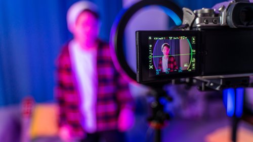Durch das Display einer Kamera sehen wir unscharf einen jungen Mann mit Mütze, der sich bei der Aufnahme eines Videos filmt. Er befindet sich in einem bläulich ausgeleuchteten Studio.