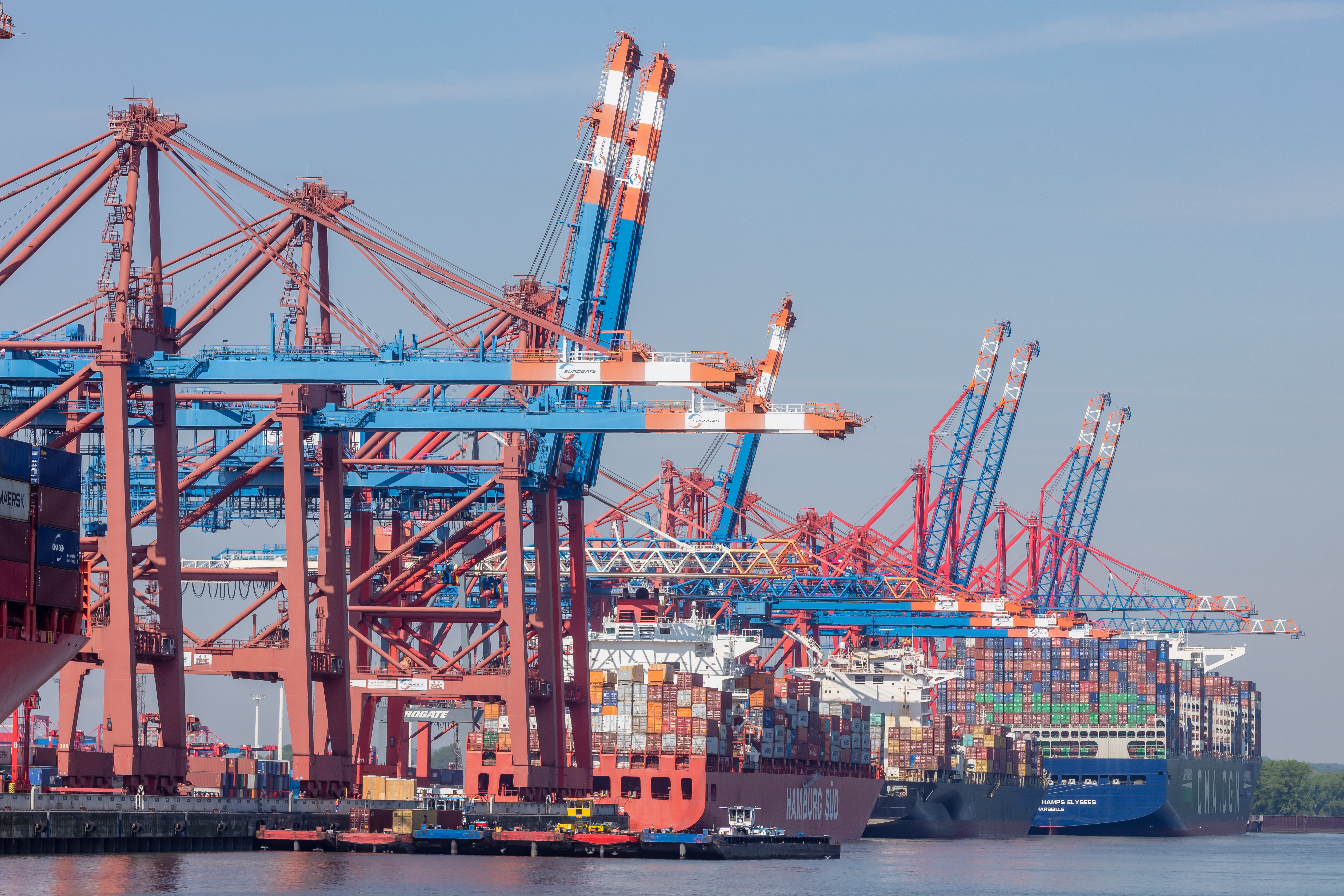 Blick auf ein Hafenterminal: Vor einer langen Reihe an Containerkränen werden zwei große Schiffe ent- oder beladen.