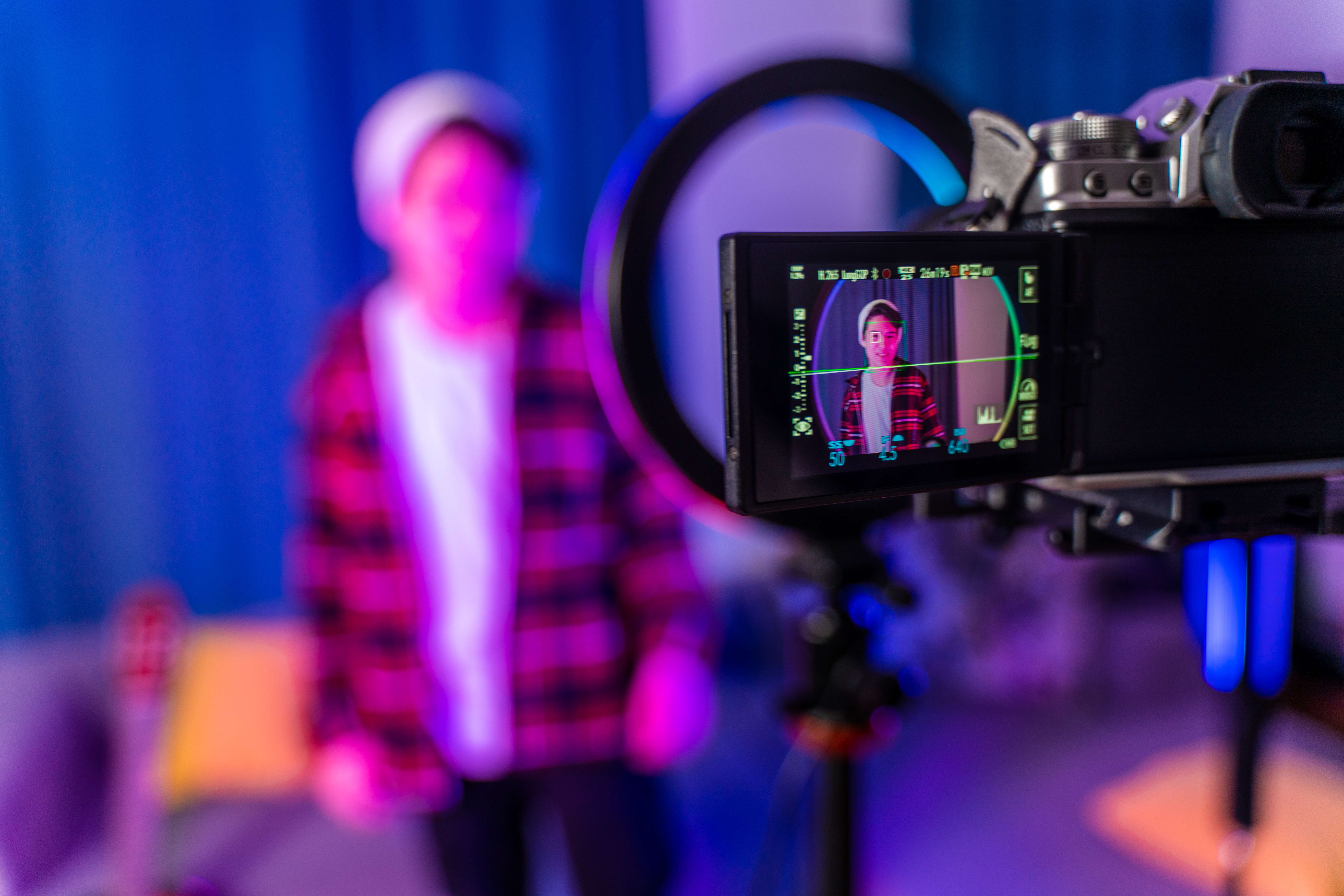 Durch das Display einer Kamera sehen wir unscharf einen jungen Mann mit Mütze, der sich bei der Aufnahme eines Videos filmt. Er befindet sich in einem bläulich ausgeleuchteten Studio.